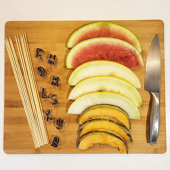 slices of melon on a cutting board with skewers, a knife, and alphabet cutters