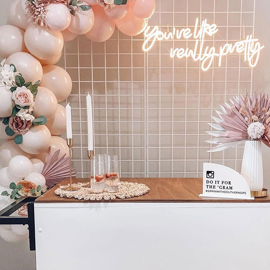 Do it for the 'Gram' custom hashtag acrylic sign on a tabletop surrounded by balloons and a neon sign