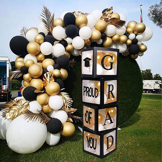 4 stacked transparent boxes filled with white and gold balloons with GRAD spelled out on the sides surrounded by balloon garland